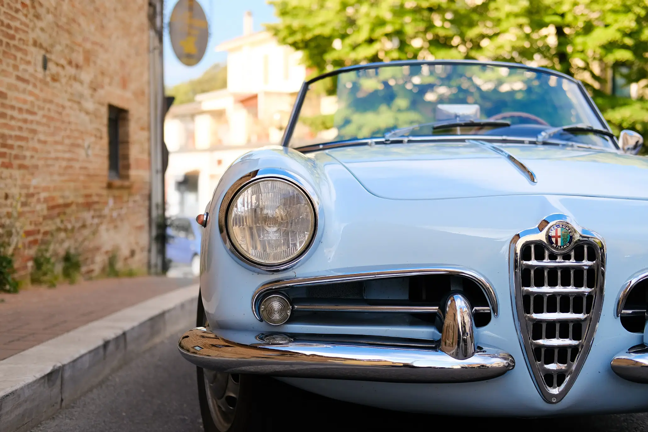 Close-up view of the chrome grille and headlight of a light blue vintage convertible parked by a brick wall.
