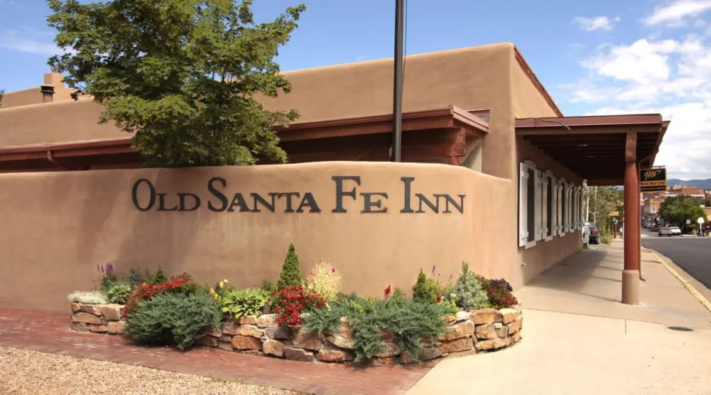 The exterior sign for the Old Santa Fe Inn on a curved adobe wall with a stone planter full of landscaping.