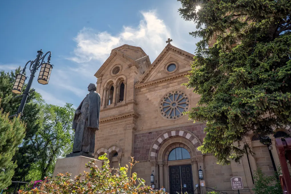 Historic stone cathedral facade featuring a rose window, a bronze statue, and tall trees against a bright blue sky.