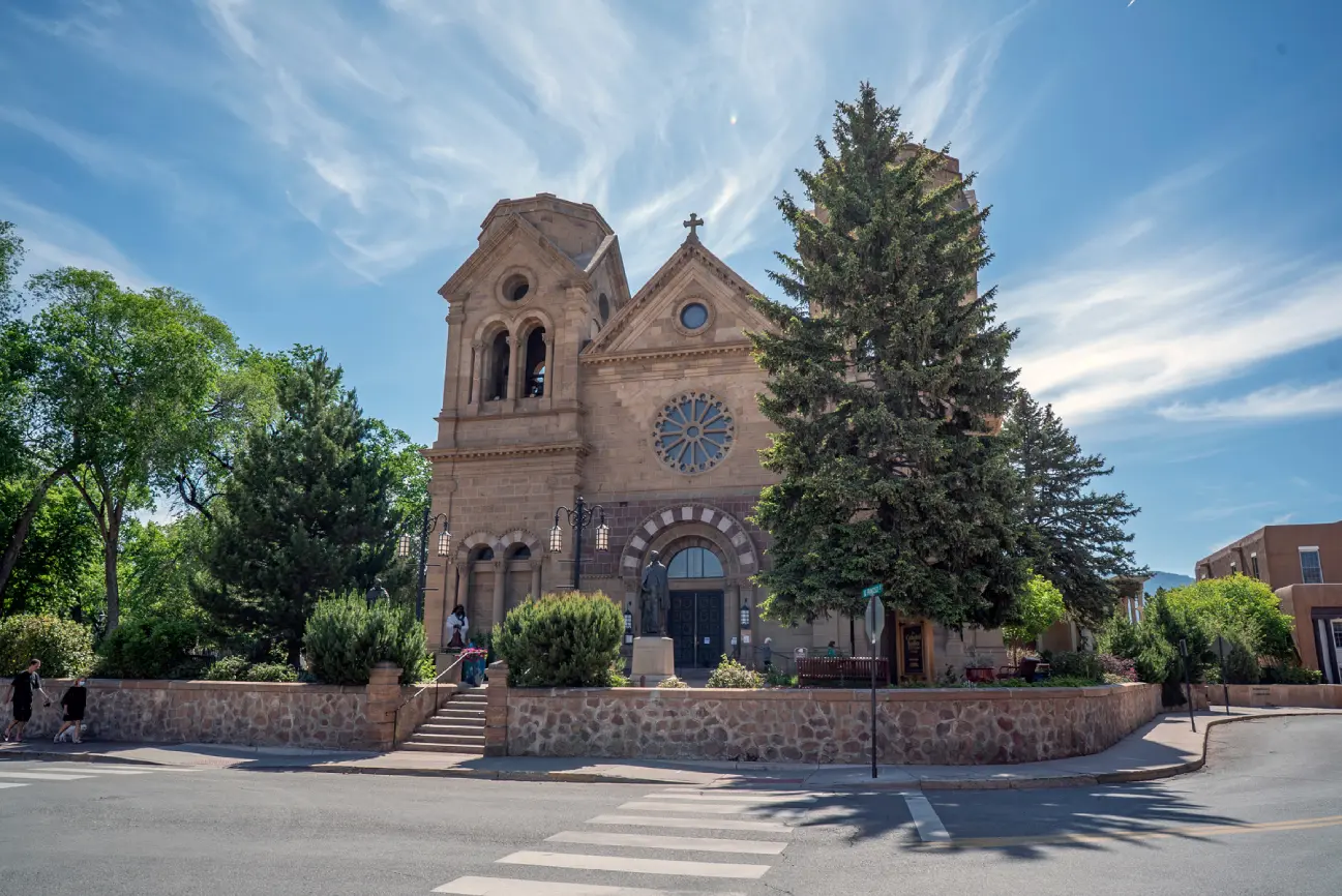 The historic stone cathedral with bell towers and a large evergreen tree is viewed from the street corner on a sunny day.