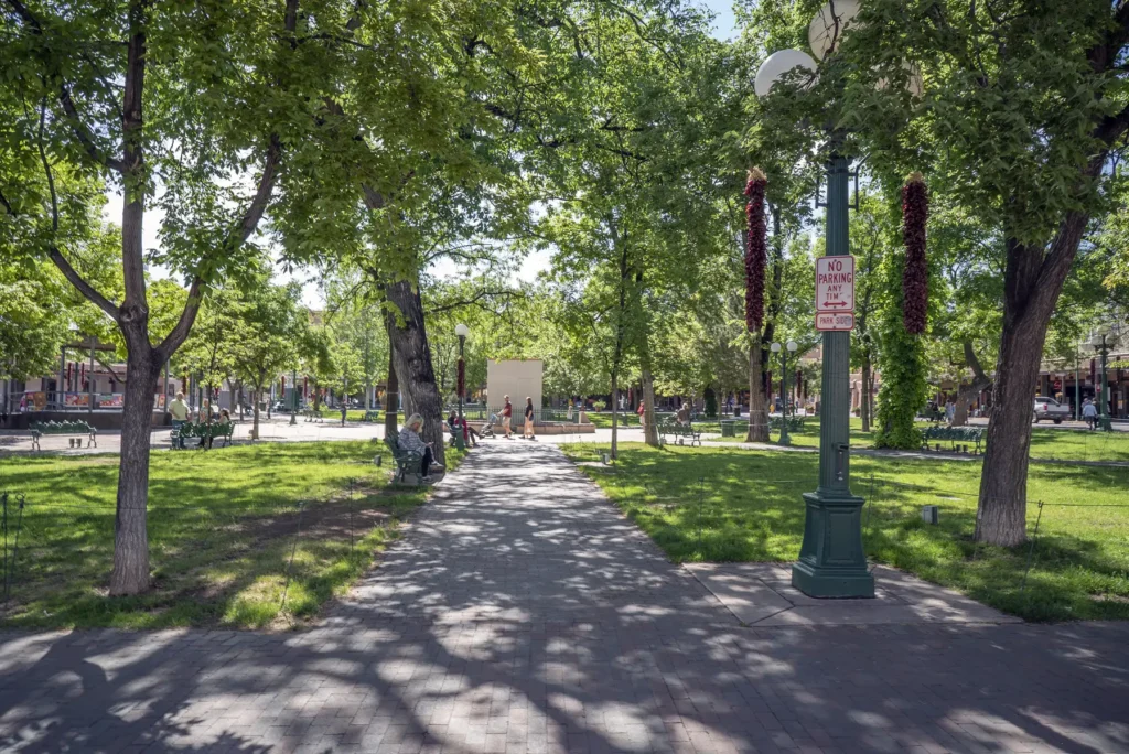 Brick pathway leading through a sunny park with tall green trees, benches, and a lamppost decorated with red ristras.