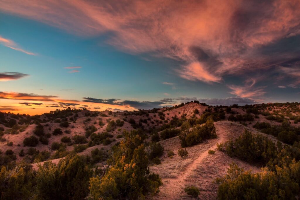 Scenic landscape of rolling desert hills covered in scrub brush under a breathtaking sky of teal and orange sunset clouds.