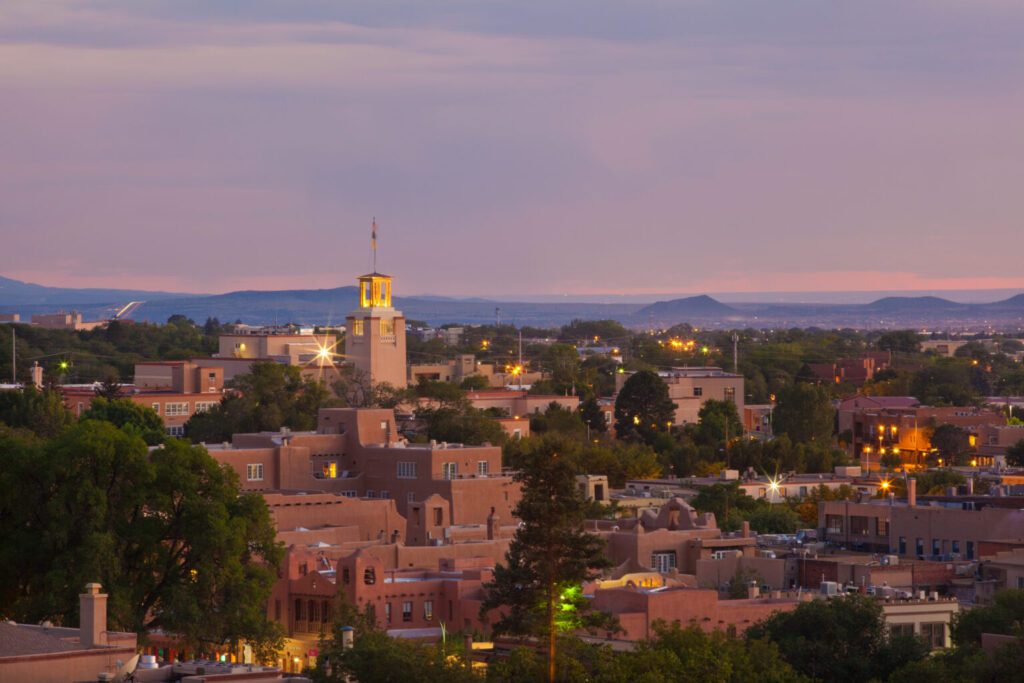 Elevated evening view of a Southwestern town with adobe buildings, trees, and a lit central tower.