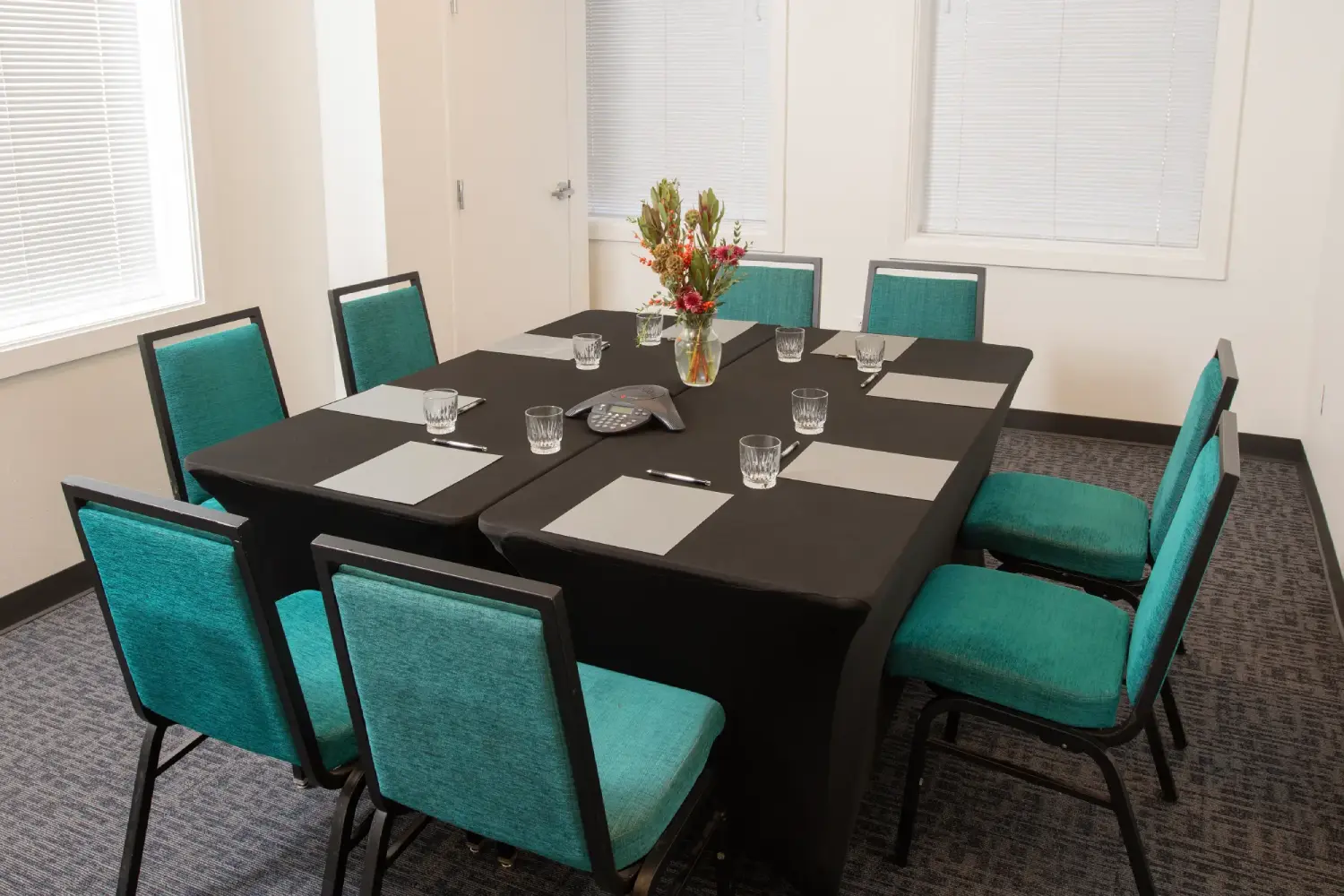 A meeting room with a black table, teal chairs, and a vase of flowers.