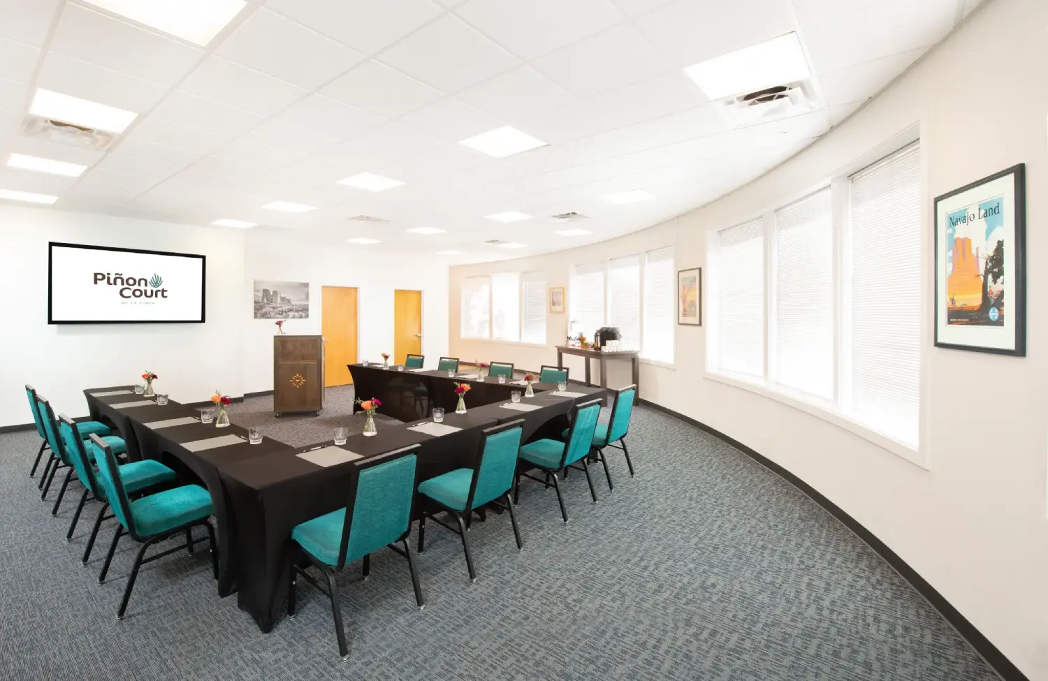 a meeting room with a long table, teal chairs, a podium, and a screen on the wall