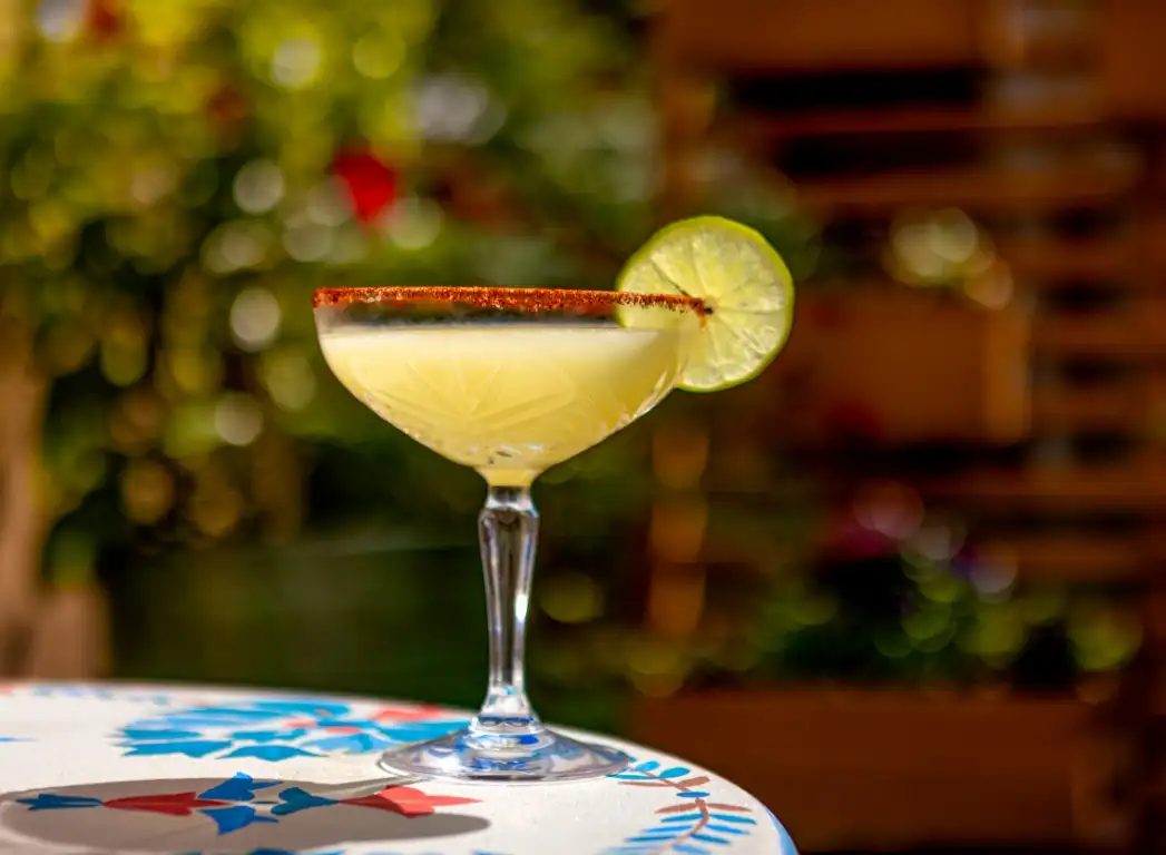 a yellow cocktail with a lime wedge and a red spice rim on a colorful table