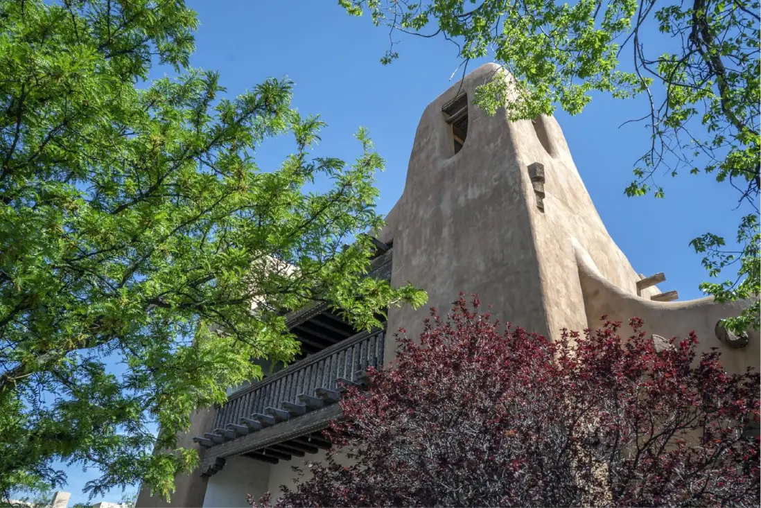 A low-angle shot of a building's exterior with a balcony, surrounded by trees and a clear blue sky.